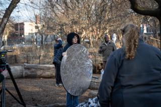 Crew member angling a large silver reflector into the scene, the unit gathered around behind