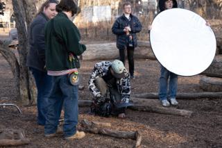 Operator crouched low over the camera while a reflector is held white-side-in above him