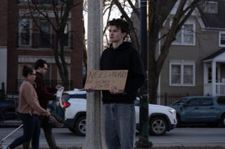 The actor in position against the light pole with his sign as passers-by cross behind him