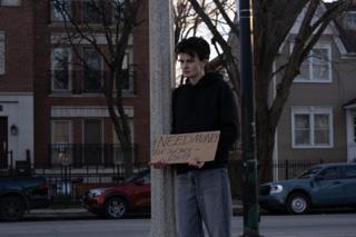 The actor holding the sign at the foot of the pole, houses and traffic behind