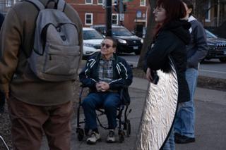 An actor waiting in his wheelchair on the sidewalk between takes, a reflector held ready beside him