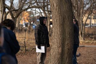 Crew member in a hooded plaid jacket holding the day's pages beside a bare tree trunk