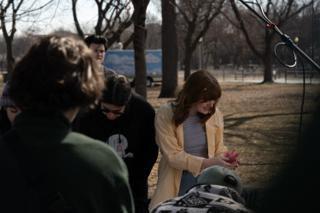 The actor turning the pink paper flower over in her hands while the crew watch from a step away