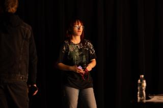 Red-haired performer in a graphic T-shirt standing under a stage light, a second figure silhouetted in the foreground