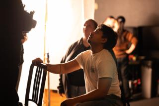 Actor seated backwards on a chair with head tilted up into the light, crew watching from behind
