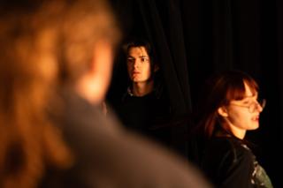 Two performers lit in warm amber between black stage curtains, a crew member out of focus in the foreground