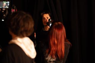 Performer holding a small camcorder up to their eye, framed between two onlookers backstage