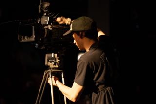 Camera operator silhouetted against warm light while framing a shot on a tripod-mounted cinema camera