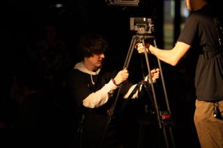 Two crew members adjusting the legs of a camera tripod on a darkened set