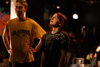 Two performers standing side by side under warm stage light, one looking upward