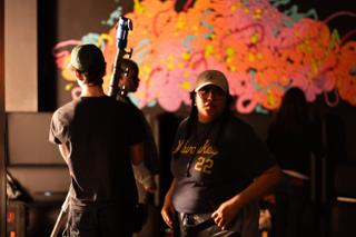 Crew member in a baseball cap carrying a light stand past a mural-painted wall while another looks on