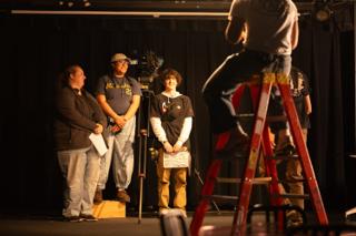 Cast and crew gathered around a step ladder on stage, one holding the clapperboard, while a grip works overhead