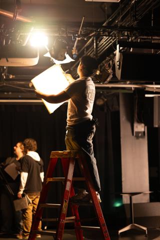 Grip on a step ladder shaping a lamp with a flag as the crew watch from below