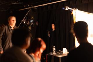 Crew watching from the shadows as a performer stands lit at the microphone beyond the camera