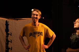 Actor grinning between takes in front of a white bounce board, a crew member at his shoulder