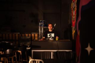Performer seated behind the bar counter on the lit set, the camera rig framing her from the dark