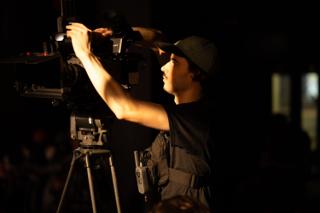 Camera operator reaching up to adjust a cinema camera mounted on a tripod