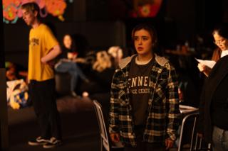 Actor standing ready on set in a plaid overshirt while the crew reset around her