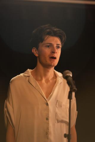 Lead actor mid-performance at the microphone, lit warm against the dark