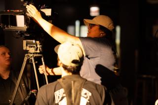 Crew member reaching over a tripod-mounted camera to adjust the monitor, seen from behind