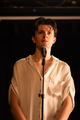 Lead actor at the microphone under a single overhead light, looking out past the camera