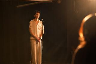 The lead actor alone at the microphone on the wide empty stage, a crew member silhouetted at the edge of frame
