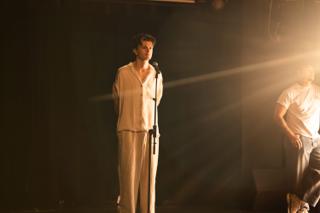 Lead actor performing in a shaft of light across the dark stage as a crew member watches from the glare