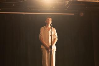 Wide of the lead actor at the microphone beneath a single hot overhead lamp