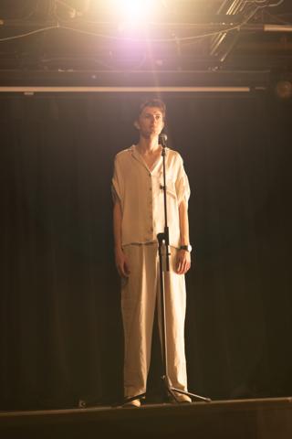 Lead actor standing full length at the microphone beneath a single hanging lamp