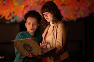 Two actors reading together from a decorated book in front of the painted mural