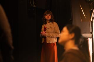 Actor standing on her mark in a tan jacket, crew heads in the foreground shadow