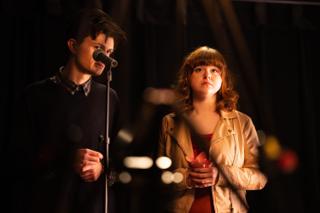 Two actors close together at the microphone, one holding a lit candle