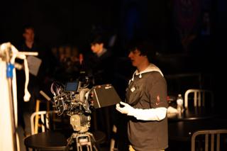Camera operator at the rig with the monitor, crew scattered through the dark room behind