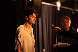 Actor standing at a microphone with eyes closed while the director holds out a script page beside them