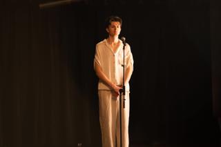 Performer in cream-coloured clothing standing alone at a microphone on a darkened stage