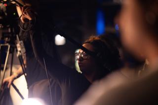 Boom operator reaching the pole out over the set, seen past a crew member in the foreground