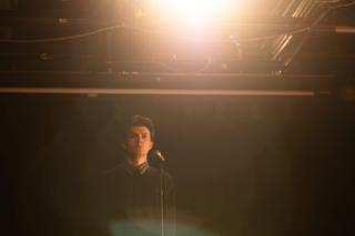 An actor alone at the microphone on the wide dark stage beneath a single flaring overhead lamp