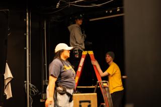Grips setting up a step ladder and stands on the darkened stage