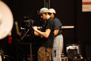 Camera operator adjusting the cinema camera on its tripod while another crew member works behind them