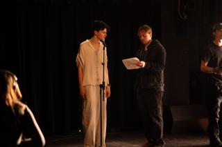 Director reading from a script to an actor at a microphone, crew visible at the edges of the stage