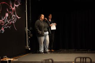 Director standing on the darkened stage with script in hand giving notes beside a performer, the mural at the edge of frame