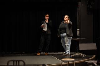 Two people standing on the empty stage against black curtains, one holding script pages mid-note