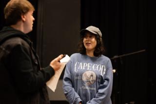Performer in conversation with a crew member holding pages, both lit warm against the black stage