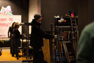 Grip pushing a loaded equipment cart past the set, mural artwork on the wall behind