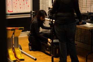 Crew member crouched over an open equipment case in the prep room, light stands stacked in the foreground