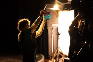 Crew member raising a clapperboard in front of a large lighting unit, a camera operator silhouetted alongside