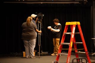 Camera team framing a shot on the darkened stage, a red step ladder standing in the foreground