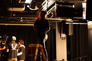 Lighting technician standing on a step ladder beneath the overhead lighting grid, warm lamps flaring around them