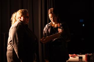 Performer in a grey coat listening as a crew member reads from a clipboard, both lit warm against the dark