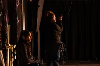 Sound crew member in headphones adjusting a cable overhead while a colleague monitors from a seat